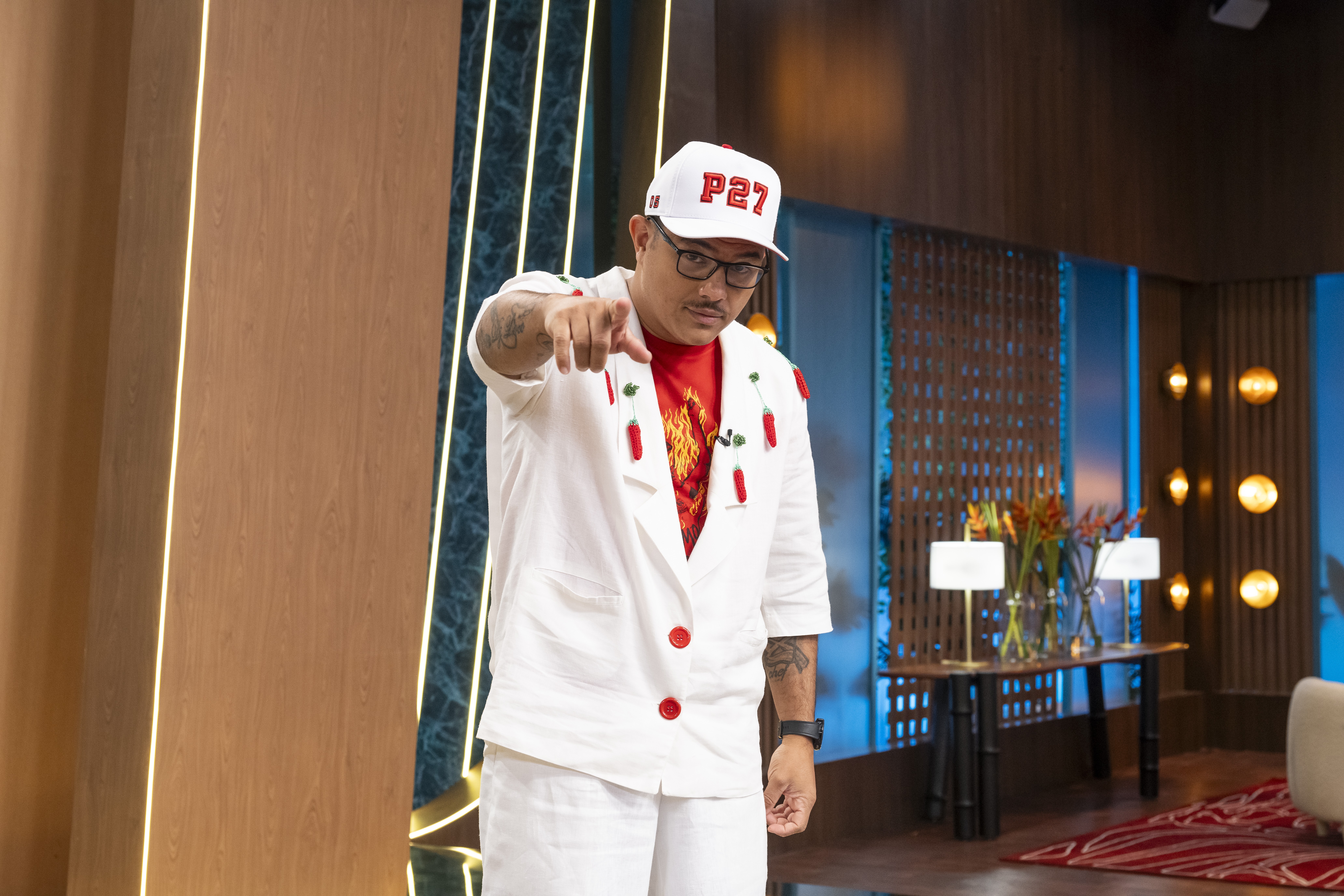 Homem de boné branco com P27 em vermelho, óculos e bigode, vestindo blazer branco com pimentas vermelhas e botões vermelhos, camiseta vermelha e calça branca, apontando para frente em um estúdio com painéis de madeira e iluminação decorativa