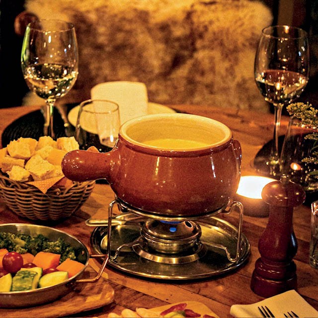 Uma panela de fondue de queijo borbulhante sobre um fogareiro, acompanhada por uma cesta de pães, uma tigela de vegetais e taças de vinho branco, em uma mesa de madeira com luz de velas