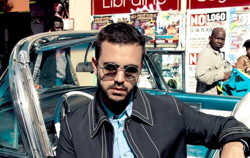 Homem branco de barba e óculos escuros redondos, vestindo camisa azul clara e jaqueta escura com costuras brancas, apoiado em um carro azul conversível. Ao fundo, pessoas e cartazes de rua