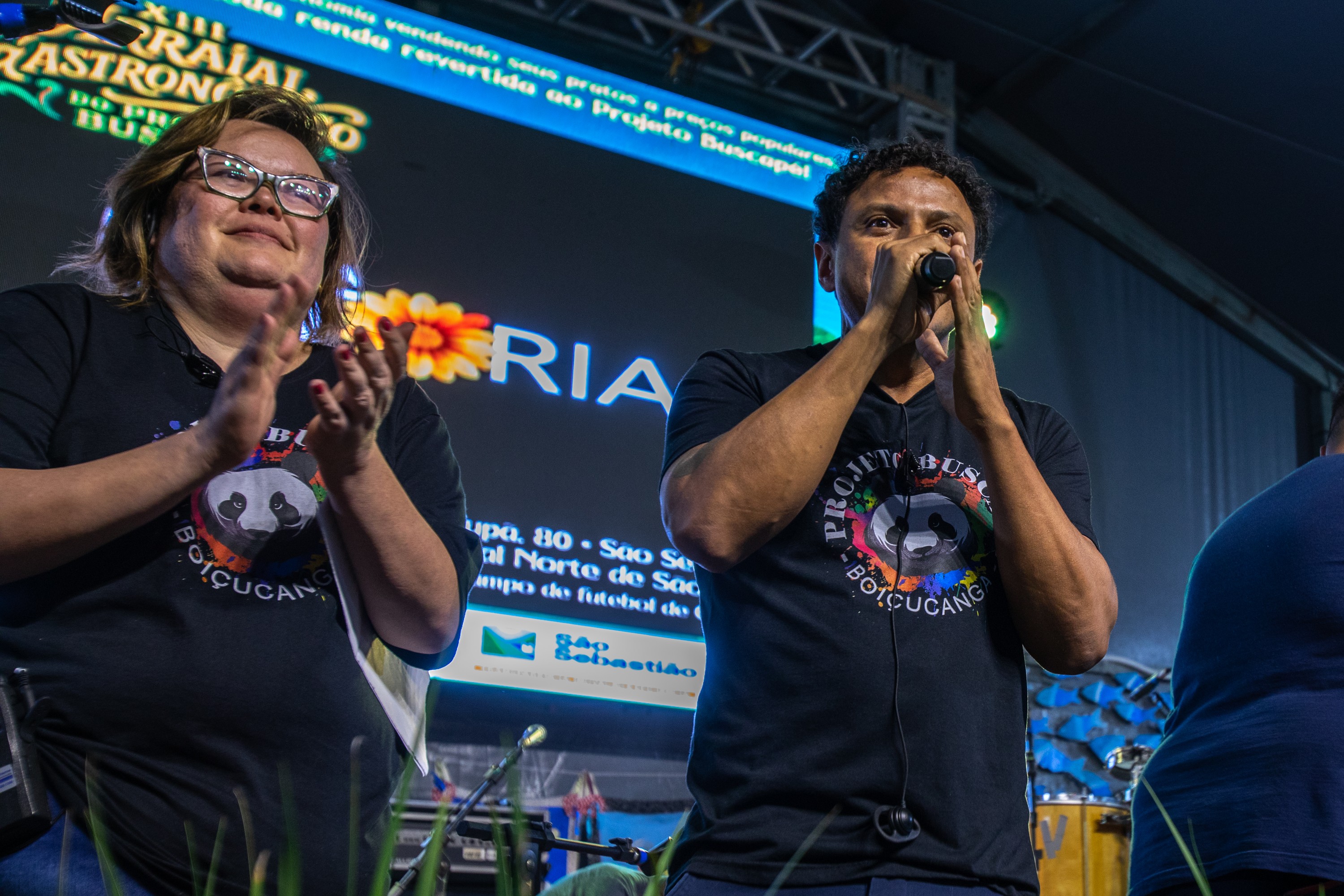 Mulher de óculos e camiseta preta com estampa de panda aplaudindo ao lado de um homem de camiseta preta com fones de ouvido, falando em um microfone. Ao fundo, um telão azul exibe texto e uma flor laranja