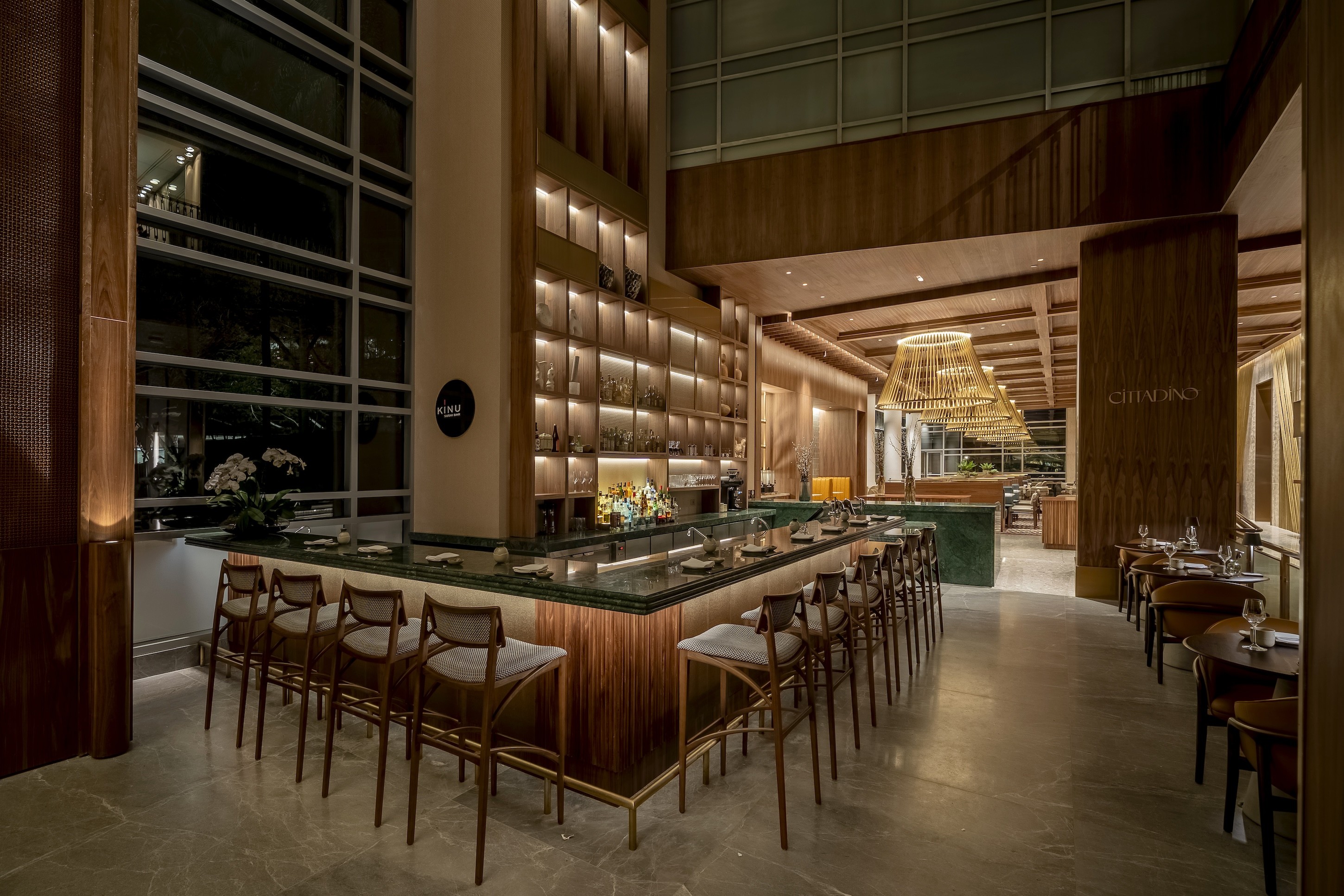 Interior de um restaurante moderno com balcão de mármore verde e banquetas de madeira, iluminado por luzes quentes. Ao fundo, prateleiras com garrafas e um grande lustre de palha. Mesas de jantar com cadeiras escuras e janelas amplas completam o ambiente sofisticado