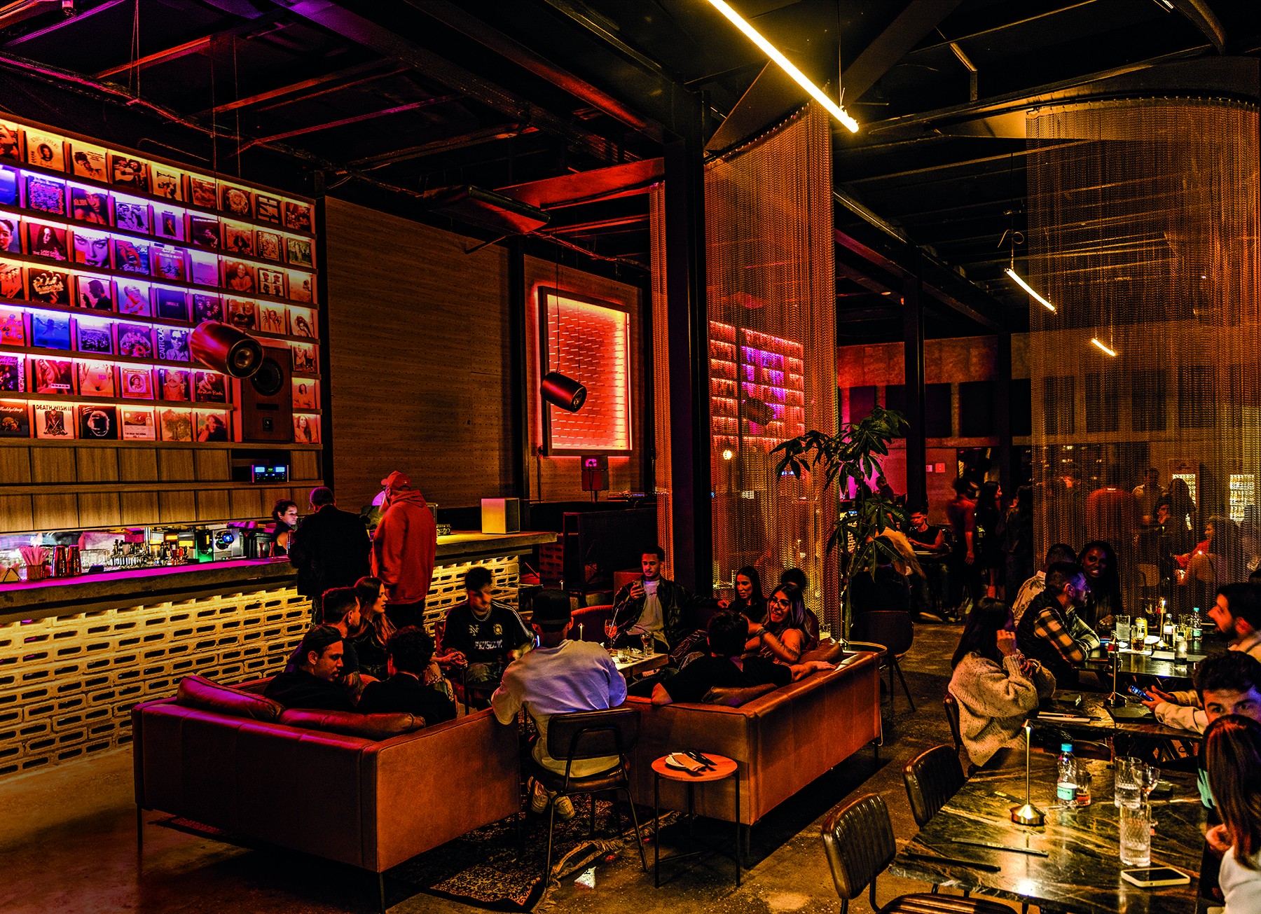 Interior de um bar ou boate com iluminação colorida e pessoas sentadas em sofás e mesas. À esquerda, uma parede com discos de vinil iluminados em tons de roxo e azul, e um balcão de bar. No centro, pessoas conversam em sofás marrons. À direita, mais mesas com clientes e uma planta grande. O ambiente é escuro com luzes neon e pendentes amarelos