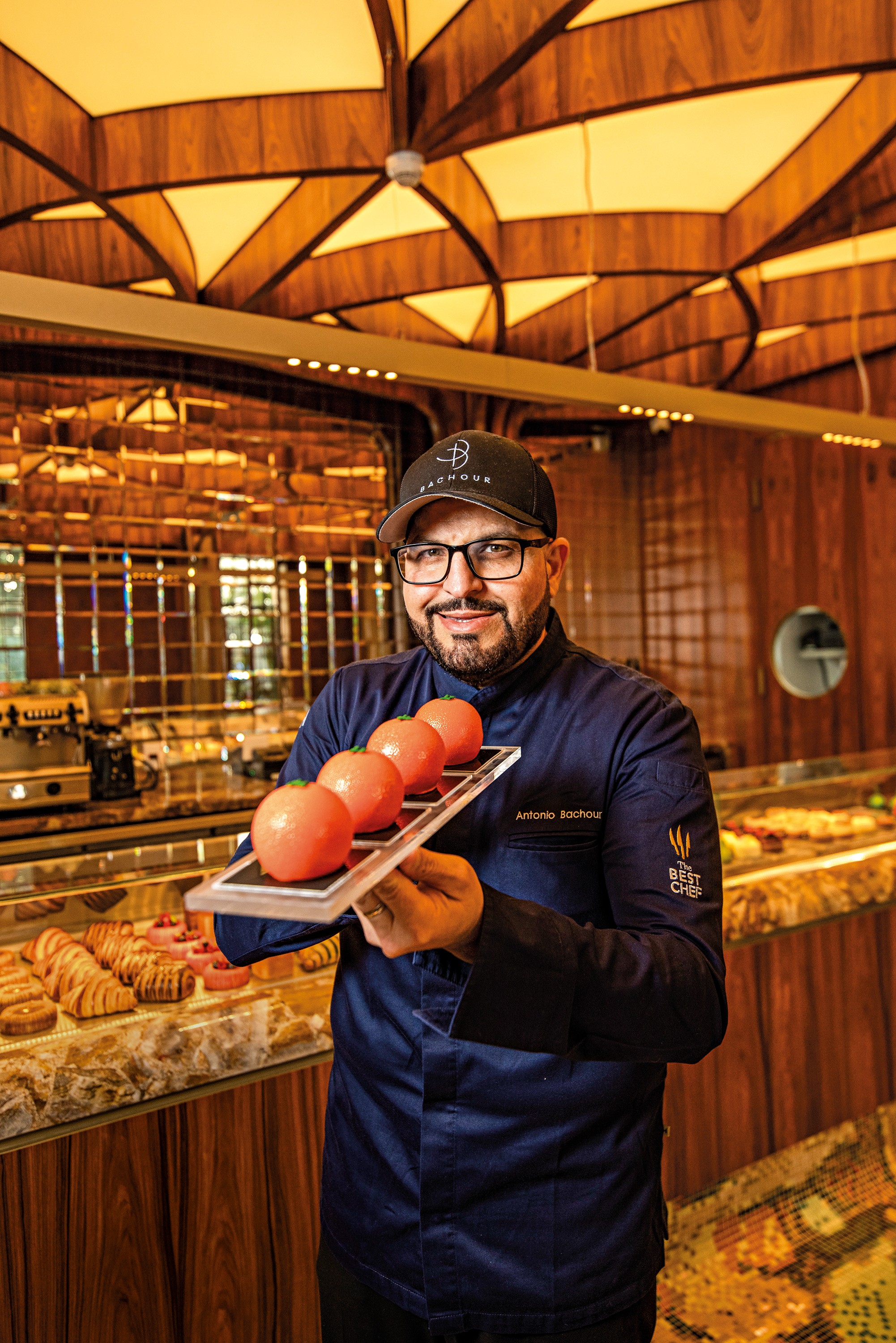 Homem de barba e óculos, vestindo uniforme de chef azul-marinho e boné preto, sorri enquanto segura uma bandeja com quatro doces redondos cor de pêssego. Ao fundo, uma confeitaria com balcões de doces e teto de madeira iluminado