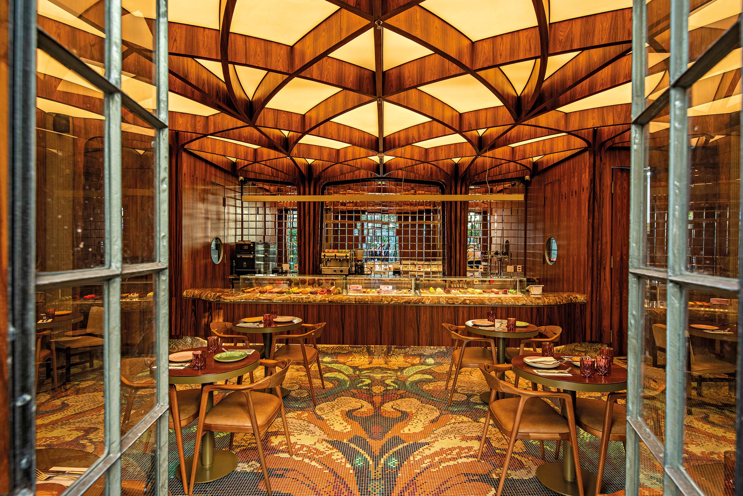 Interior de um restaurante luxuoso com teto abobadado de madeira e iluminação amarela suave. Ao centro, um balcão de buffet com alimentos. Mesas redondas de madeira com cadeiras de couro marrom estão dispostas sobre um tapete estampado com motivos de pavão. As paredes são de madeira escura, e grandes janelas de vidro emolduram a cena