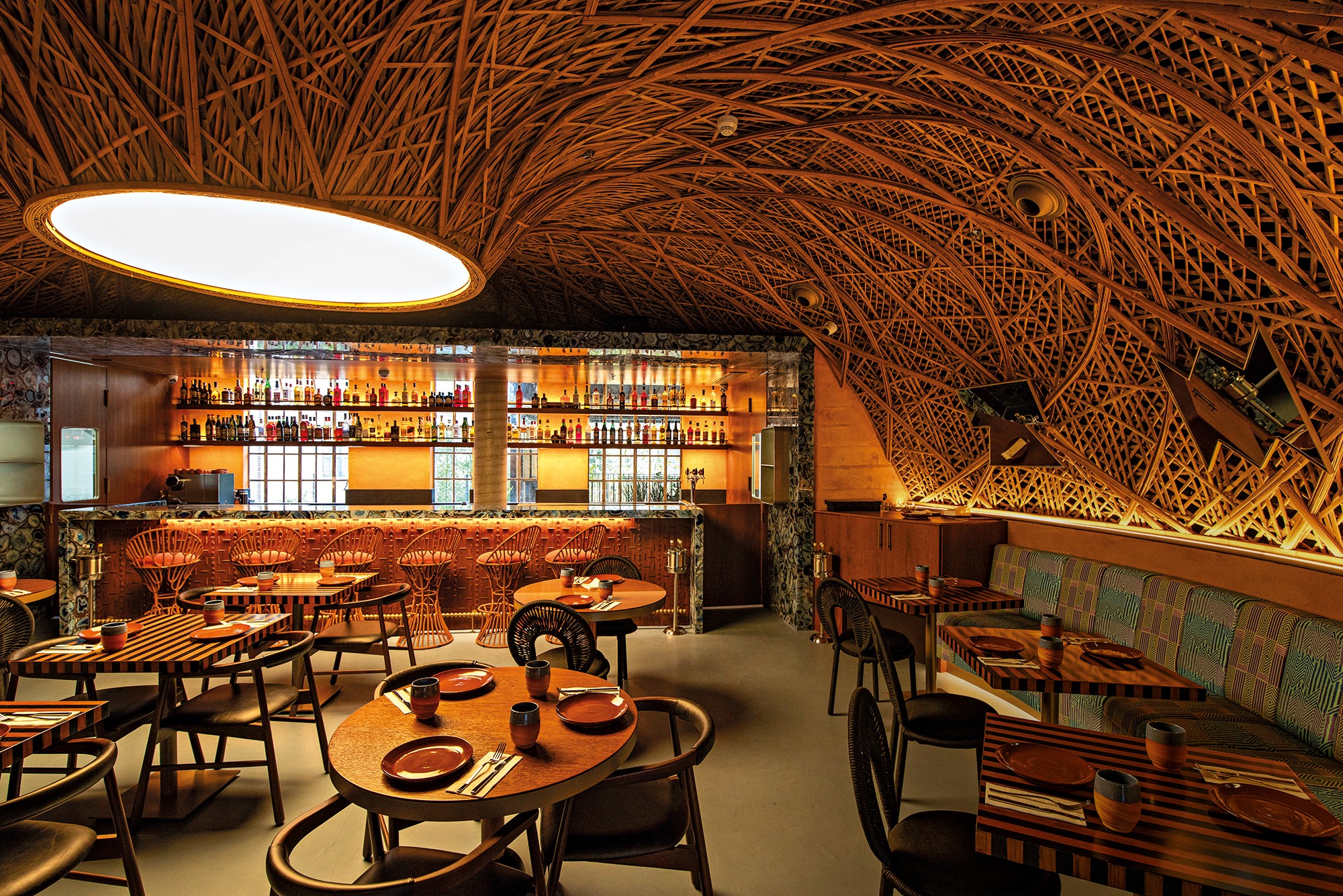 Interior de um bar ou restaurante com teto abobadado de madeira entrelaçada. À esquerda, um balcão com garrafas iluminadas e banquetas de vime. Mesas de madeira com pratos e talheres estão dispostas pelo salão, algumas com cadeiras escuras e outras com um longo sofá estampado. A iluminação é suave, criando um ambiente acolhedor