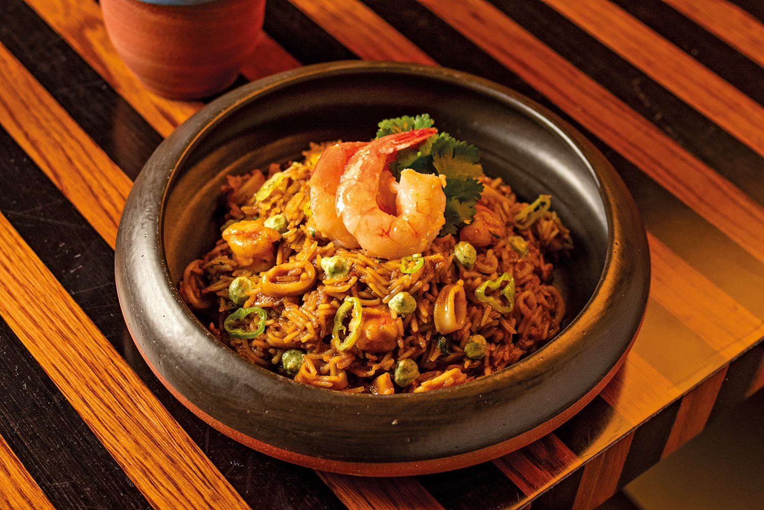 Arroz com frutos do mar, camarões, lulas, ervilhas e pimentões verdes fatiados, servido em uma tigela de cerâmica escura sobre uma mesa de madeira com listras diagonais marrons e laranjas