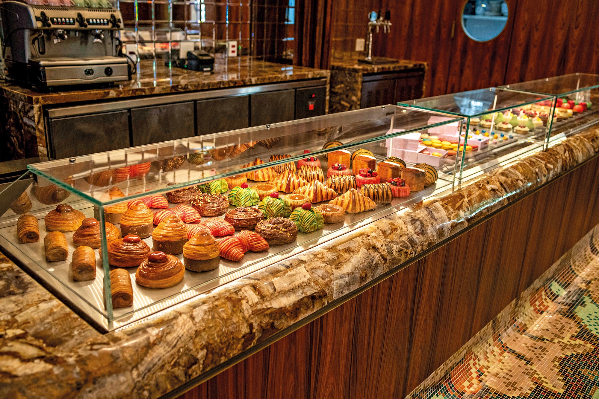 Exibição de doces e pães em uma vitrine de vidro iluminada, com uma variedade de croissants, bolos e tortas coloridas. Ao fundo, uma máquina de café expresso e balcões de mármore.