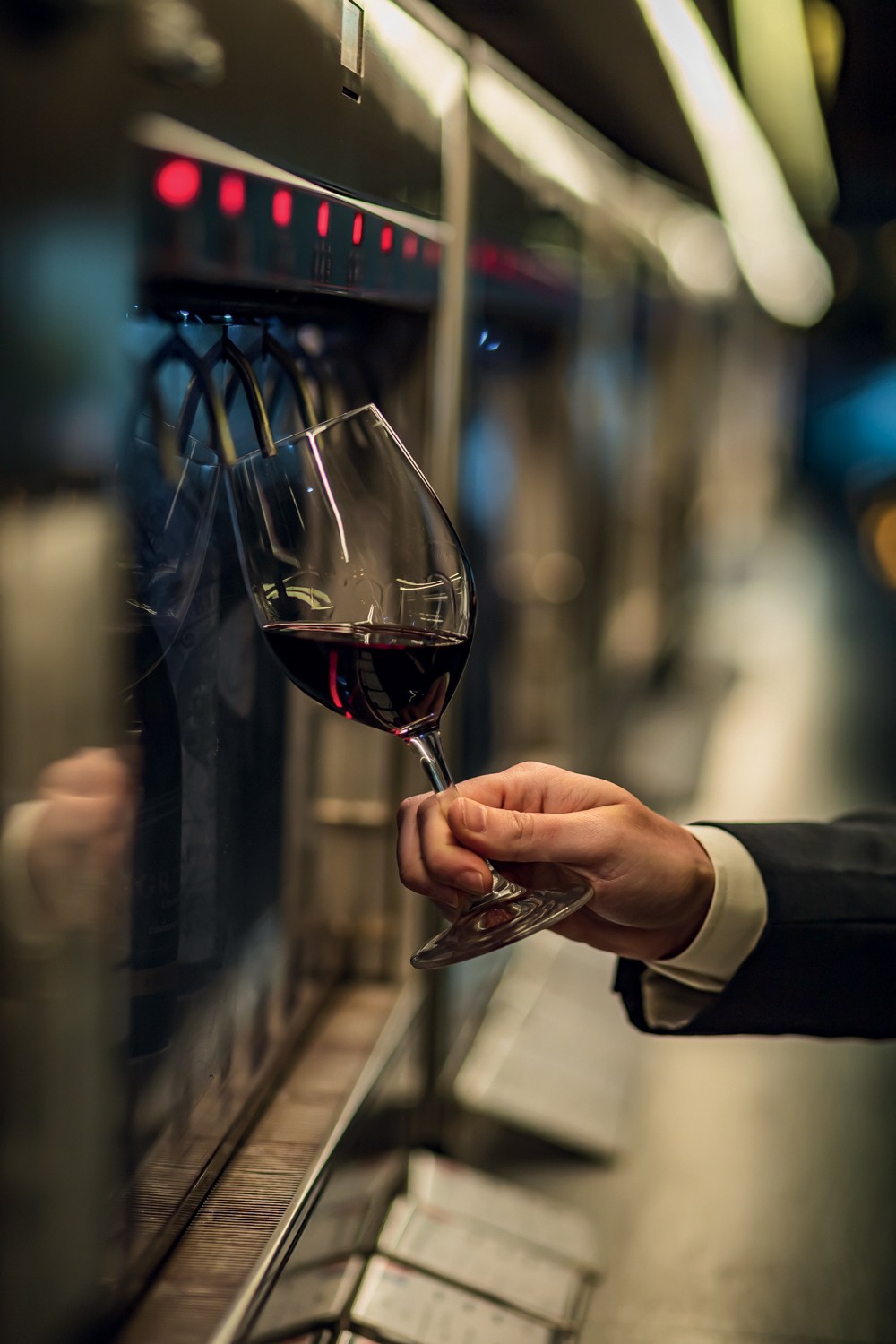 Mão masculina segurando uma taça de vinho tinto, sendo servida por uma máquina dispensadora com luzes vermelhas em um ambiente escuro