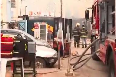 Bombeiros com uniformes de proteção e capacetes, um deles segurando uma mangueira, próximos a um caminhão vermelho de bombeiros e um carro prateado danificado. Ao fundo, fumaça escura e um estabelecimento comercial com cartazes de cerveja, indicando um incêndio em área urbana