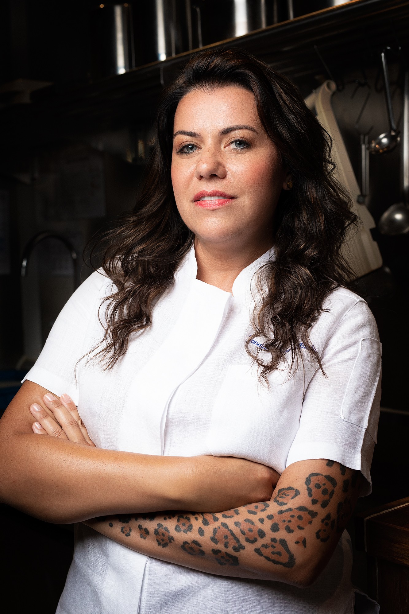 Mulher de pele morena, cabelos castanhos escuros e ondulados, vestindo dólmã branco, braços cruzados, sorri levemente. Ela tem uma tatuagem de onça no antebraço direito e está em uma cozinha com fundo escuro e utensílios metálicos pendurados