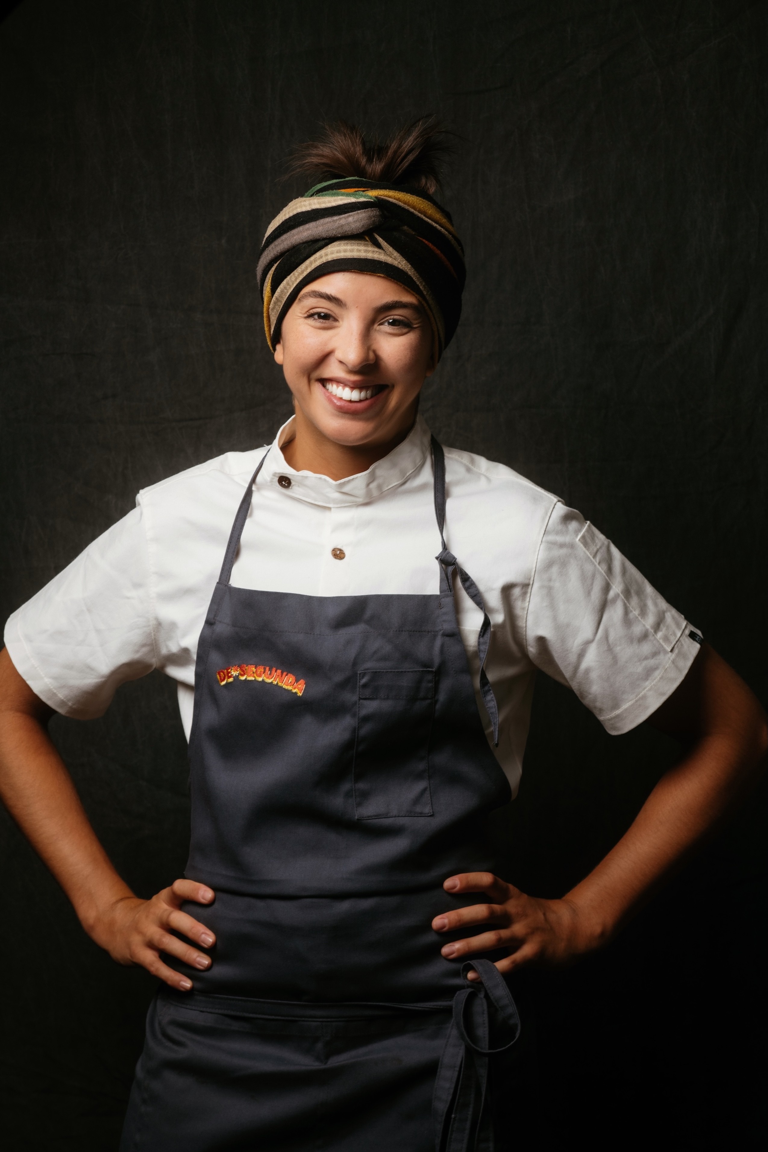 Mulher sorridente, pele morena, vestindo dólmã branco e avental cinza com o logo DESENROLA em laranja. Ela usa um turbante listrado em tons de preto, bege e verde, e tem as mãos na cintura, em fundo escuro
