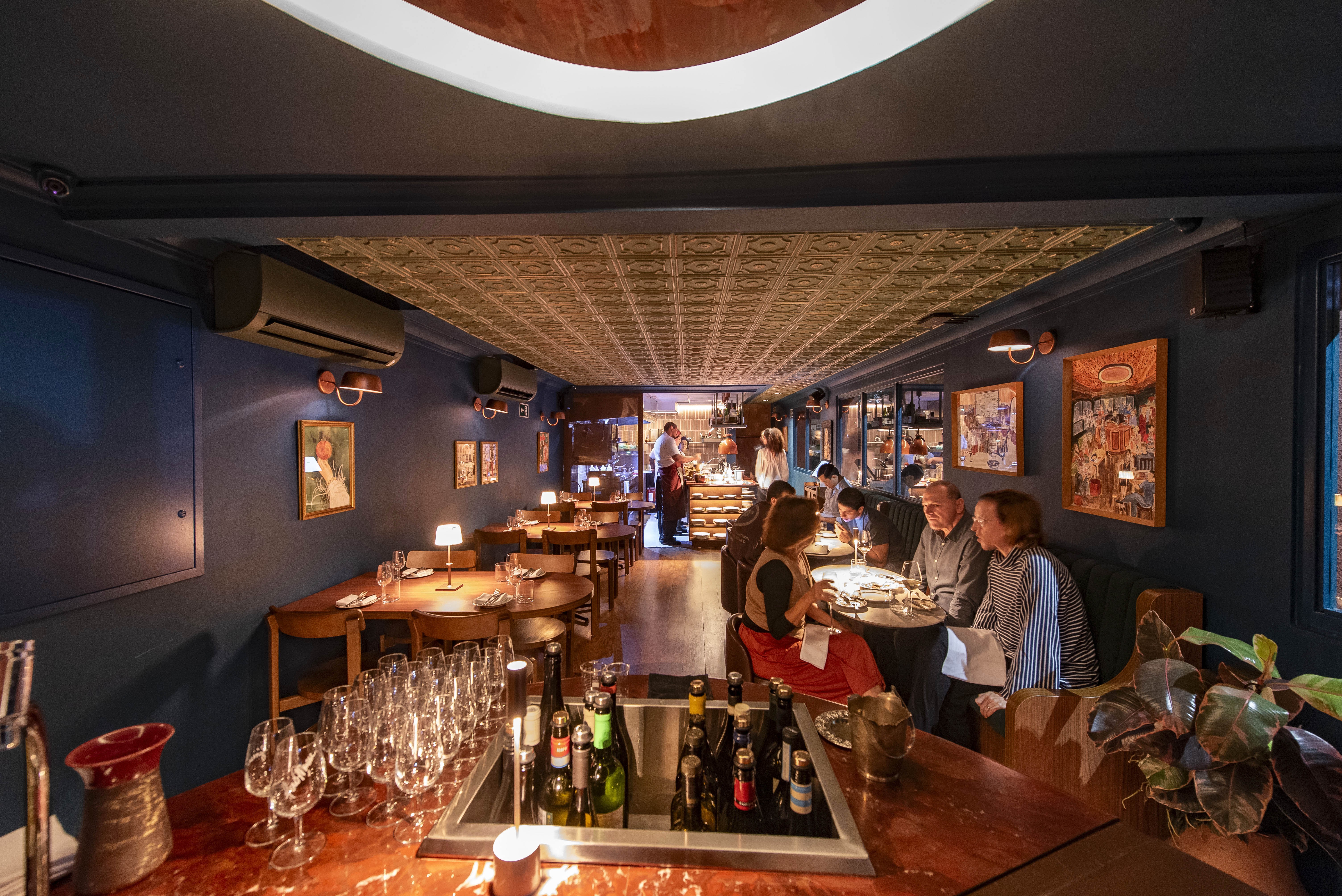 Interior de restaurante com teto dourado texturizado e paredes azul-marinho. Clientes sentados em mesas de madeira escuras, alguns conversando e outros olhando para seus pratos. Um bar com garrafas de vinho e taças vazias ocupa o primeiro plano.