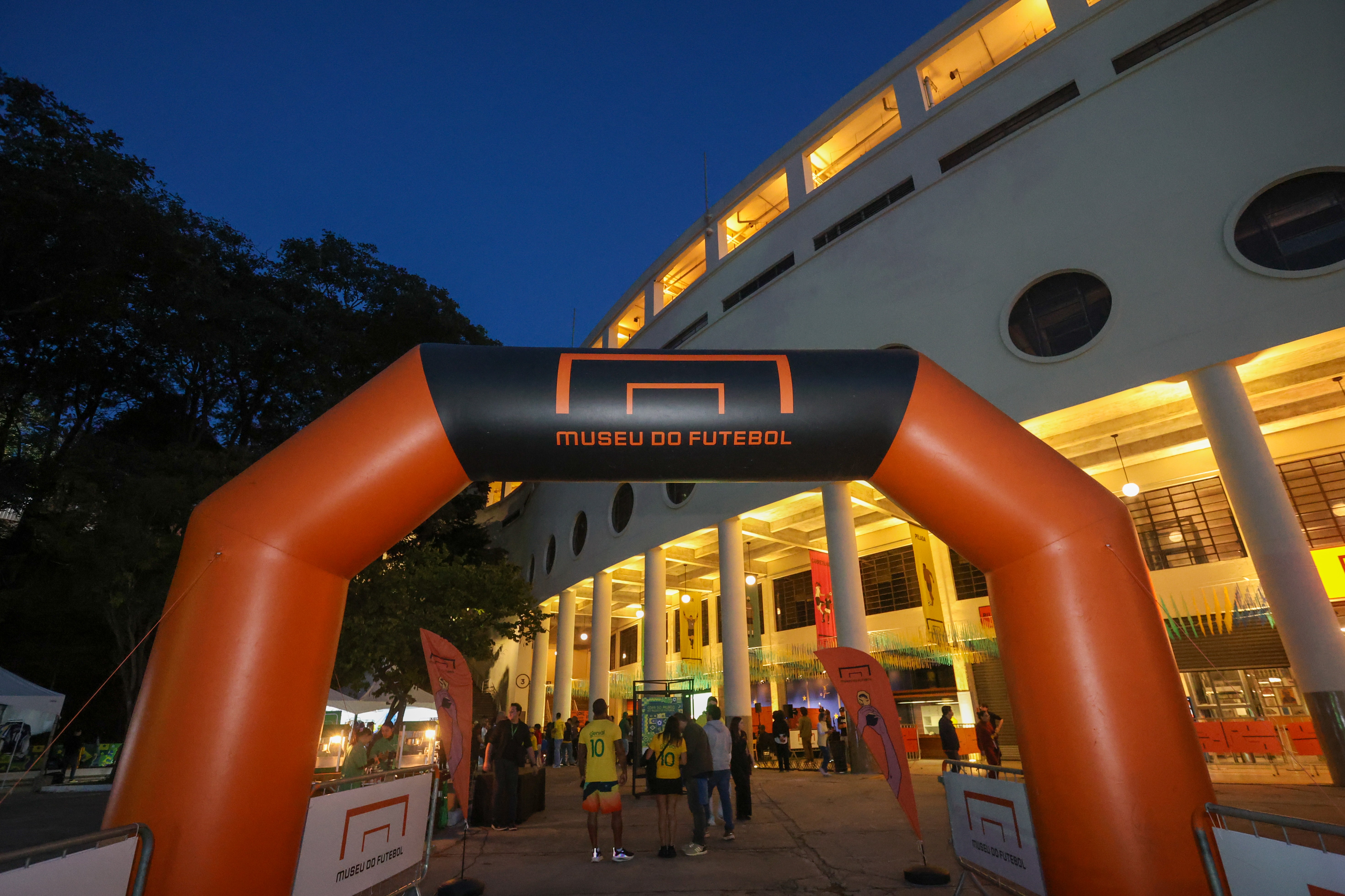Arco inflável laranja e preto com a inscrição MUSEU DO FUTEBOL e um gol estilizado, em frente a um edifício iluminado com colunas brancas, sob um céu azul escuro. Pessoas circulam na entrada do museu.