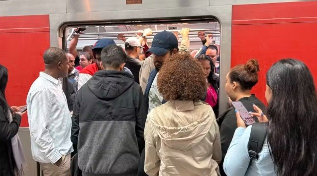 Pessoas aglomeradas na porta de um trem vermelho, algumas dentro e outras tentando entrar, indicando superlotação. Um homem de camisa listrada e outro de moletom cinza e preto estão de costas, enquanto uma mulher de cabelo cacheado e outra de cabelo liso observam a cena. Mãos seguram as barras internas do trem