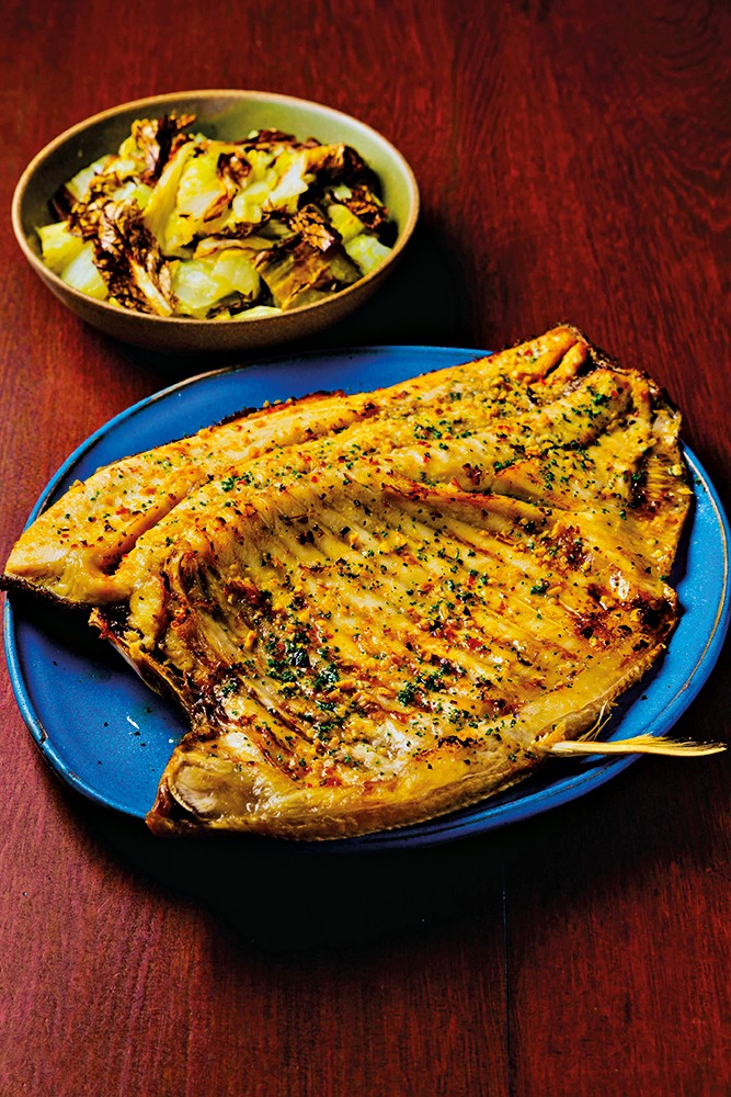 Peixe grelhado inteiro, dourado e com ervas picadas, servido em prato azul, acompanhado de repolho refogado em tigela marrom, sobre mesa de madeira escura