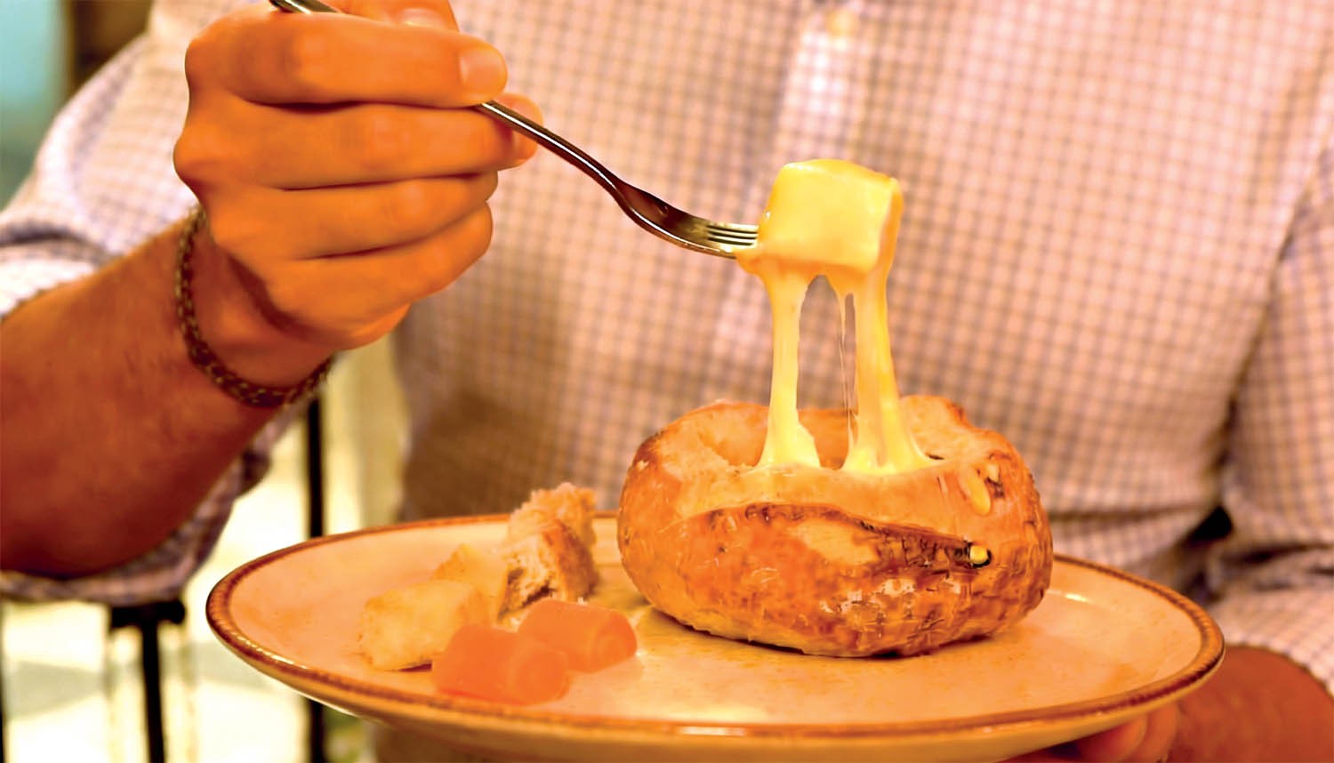 Pessoa com camisa xadrez segurando um garfo com um pedaço de pão mergulhado em fondue de queijo, que escorre do pão para dentro de um pão italiano oco, servido em um prato com outros pedaços de pão e frutas cristalizadas