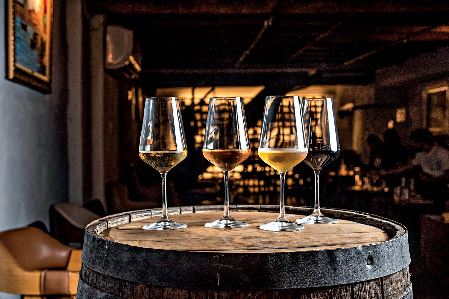 Quatro taças de vinho com diferentes tonalidades de líquido, do claro ao escuro, sobre um barril de madeira em um ambiente escuro de bar com pessoas desfocadas ao fundo