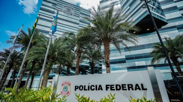 Fachada do prédio da Polícia Federal, com o letreiro POLÍCIA FEDERAL em destaque, o brasão da instituição, e várias palmeiras em frente. Ao fundo, um edifício moderno com janelas espelhadas em tons de azul e cinza, e mastros com bandeiras do Brasil e de um estado. O céu está azul com algumas nuvens