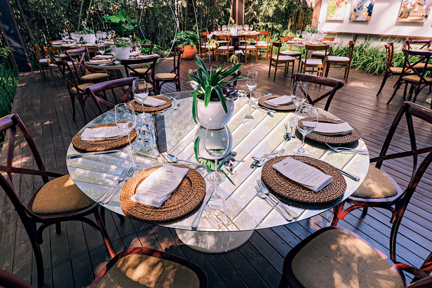 Mesa redonda de vidro com oito lugares, posta para refeição ao ar livre. Cada lugar tem um sousplat de palha, guardanapo branco com menu, talheres e taças. Um vaso branco com planta verde centraliza a mesa. O chão é de madeira e há outras mesas ao fundo, cercadas por vegetação