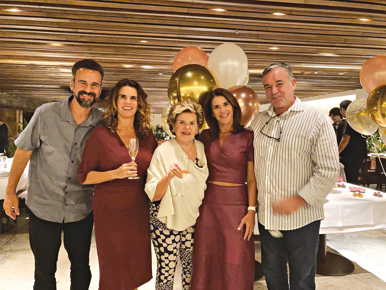 Cinco pessoas sorrindo para a câmera em um evento, com balões dourados e rosê ao fundo. Um homem de barba e camisa cinza, duas mulheres de vestido vinho, uma senhora de blusa creme e calça estampada, e um homem de camisa listrada e óculos pendurados