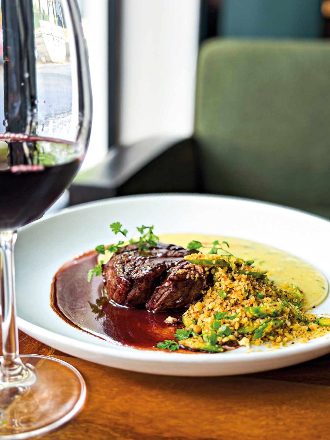 Prato gourmet com medalhões de carne ao molho escuro, purê amarelo e farofa verde com ervas frescas, ao lado de uma taça de vinho tinto, sobre uma mesa de madeira