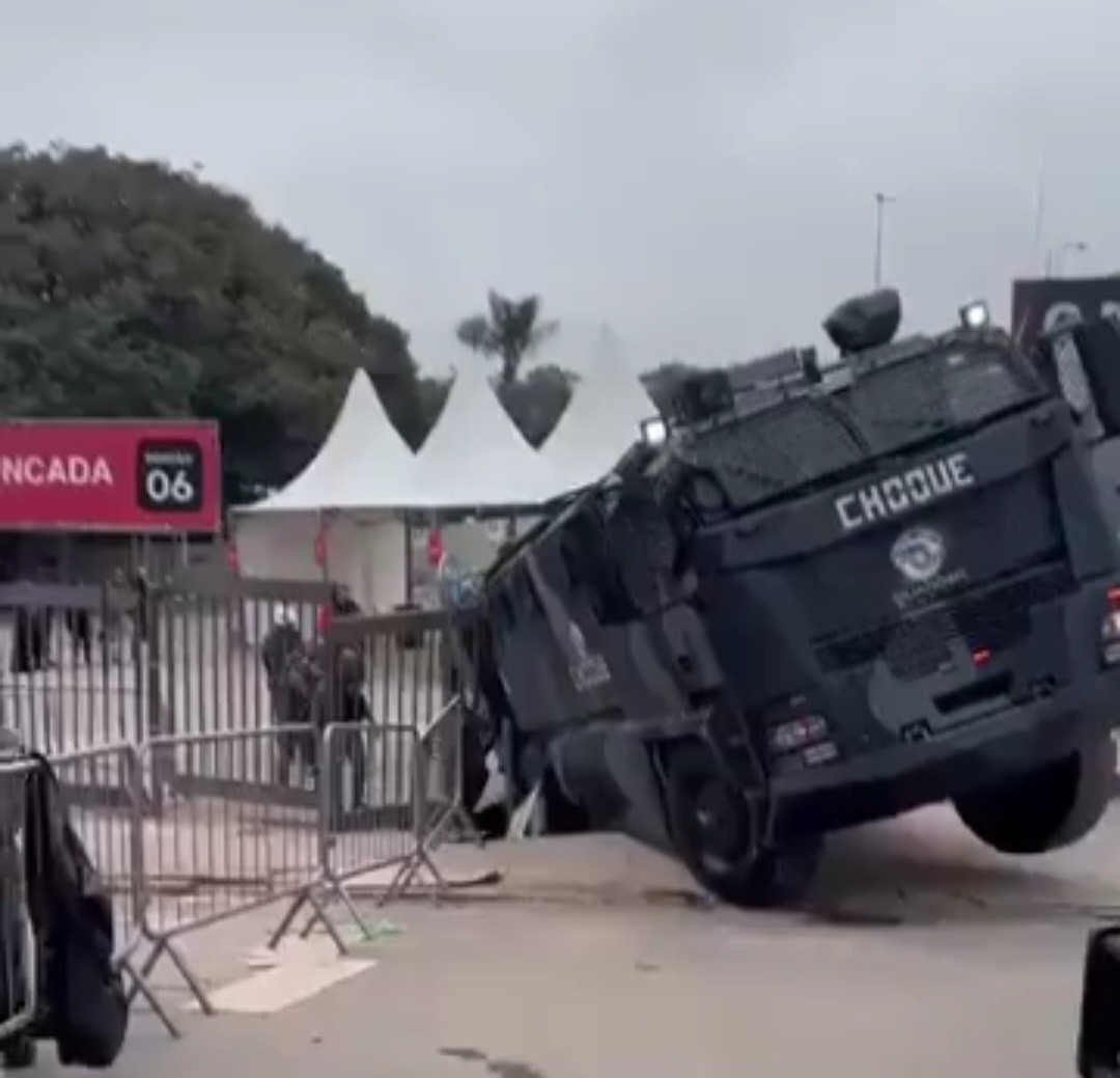 Veículo blindado da polícia, com a palavra CHOQUE na lateral, tombado sobre o lado direito em uma área externa com grades de contenção e tendas brancas ao fundo, sob céu nublado