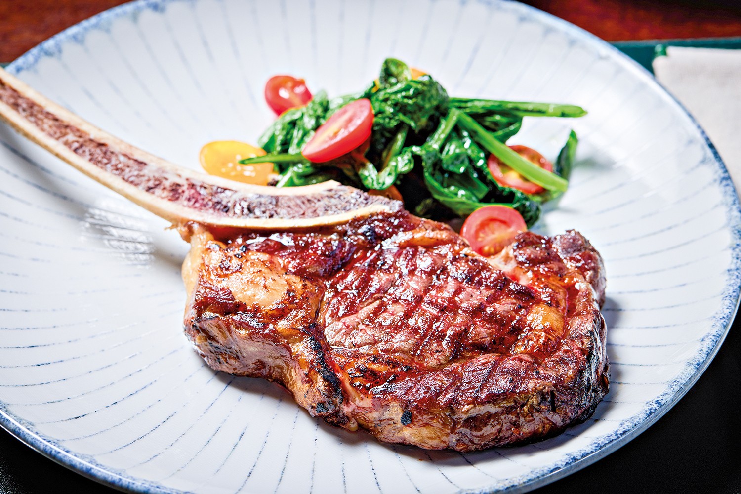 Bife tomahawk grelhado com osso, suculento e com marcas de grelha, servido em prato branco com borda azul e linhas finas. Ao lado, um acompanhamento de folhas verdes escuras, tomates cereja cortados ao meio e pimentões amarelos