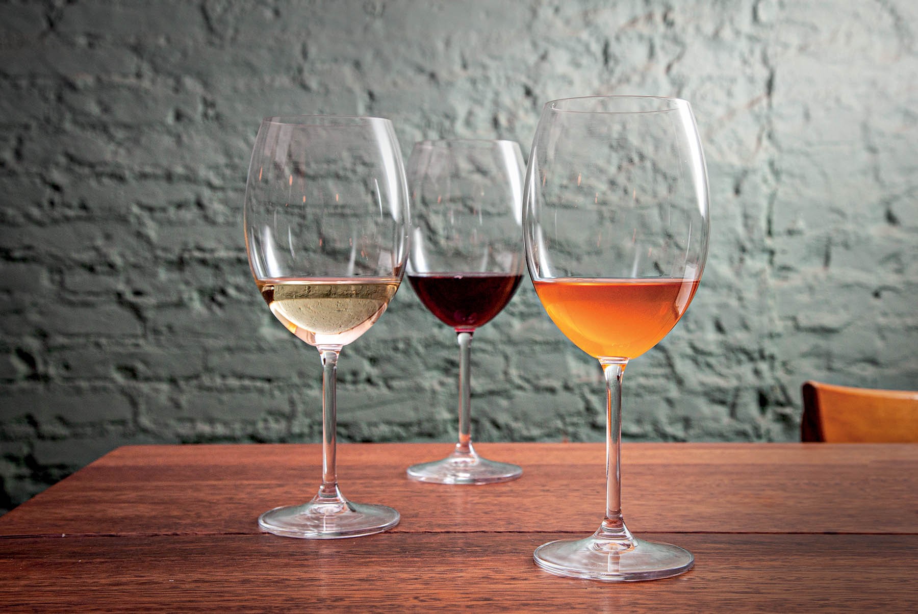 Três taças de vinho em uma mesa de madeira escura, com uma parede de tijolos cinza ao fundo. Da esquerda para a direita, as taças contêm vinho branco, tinto e rosé.