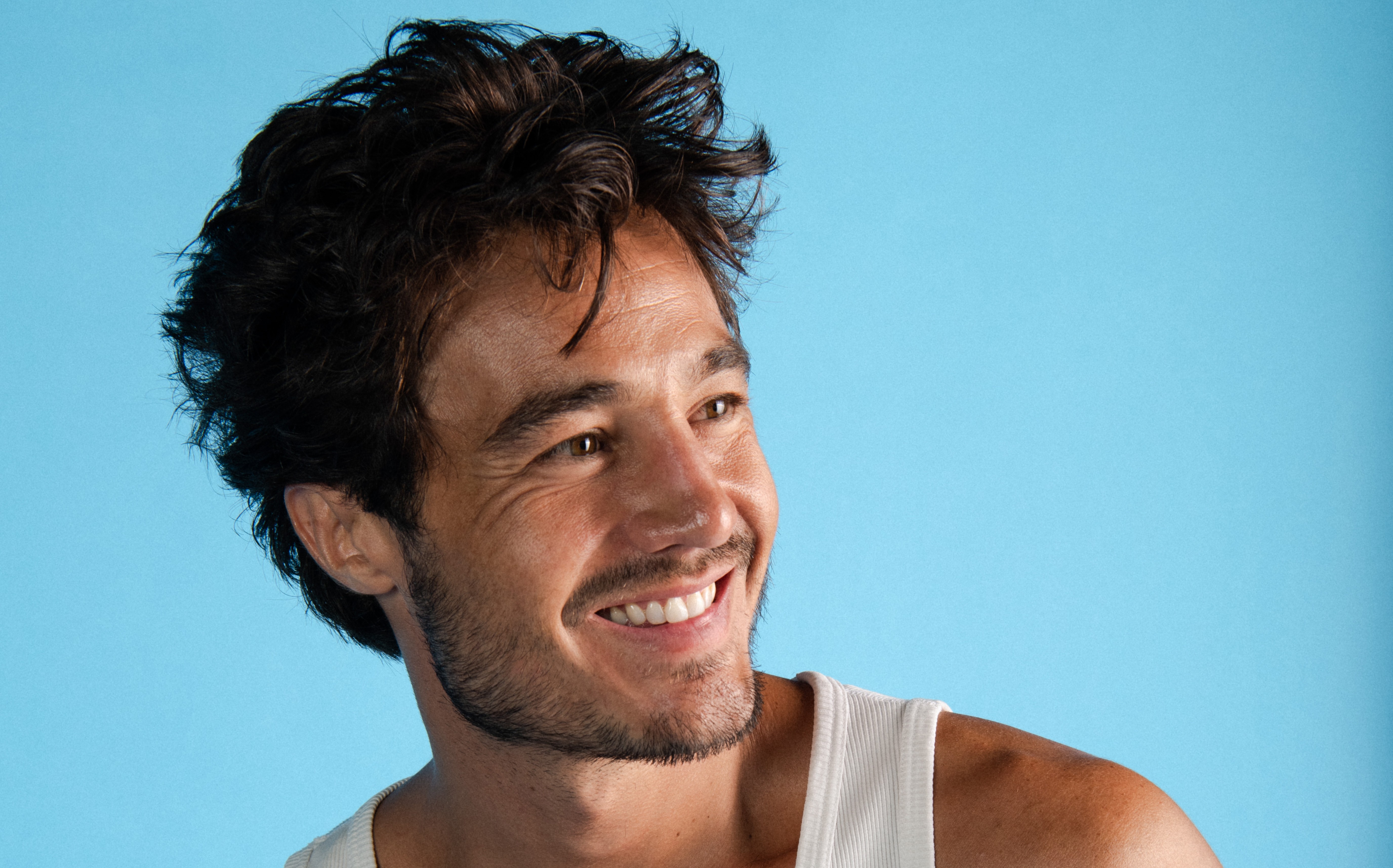 Homem de pele bronzeada, cabelo castanho escuro e barba rala, sorrindo e olhando para a direita, vestindo uma regata branca, em fundo azul claro
