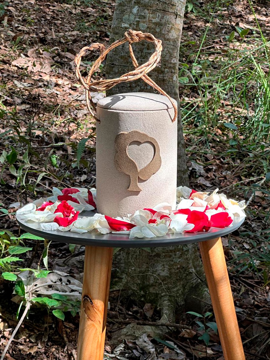 Urna funerária bege com alça de cipó e um coração vazado em relevo, sobre uma mesa de madeira com pétalas de rosas brancas e vermelhas, em meio a uma floresta