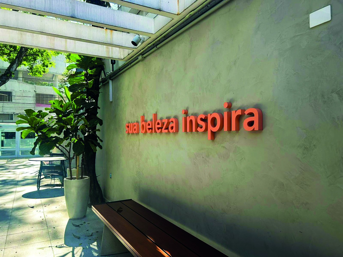 Parede verde-acinzentada com a frase sua beleza inspira em letras 3D laranja. À esquerda, um vaso branco com planta verde, uma mesa redonda e cadeiras. Um banco de madeira escura está em primeiro plano, à direita. O ambiente é externo, com luz solar e um pergolado branco no topo