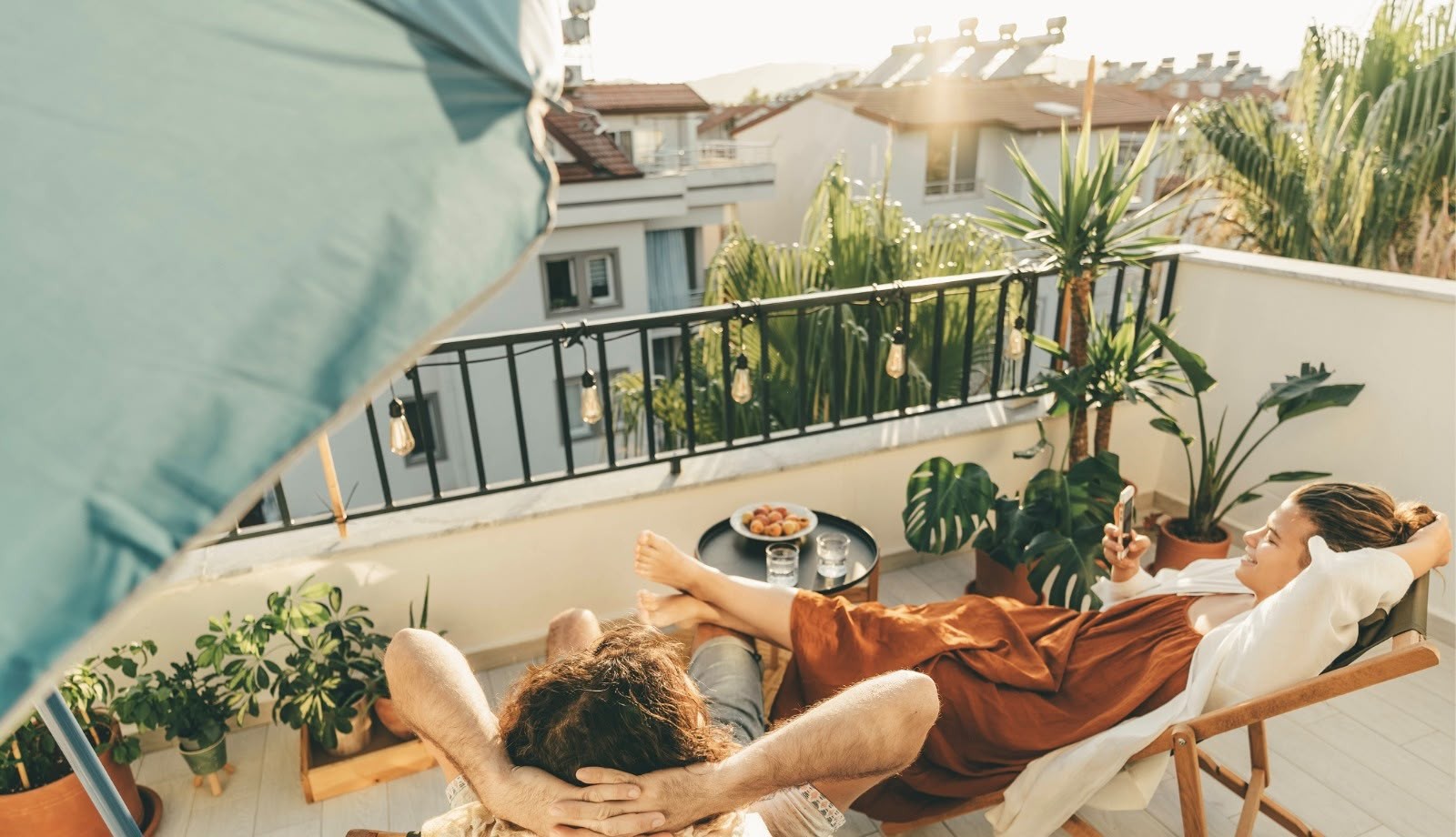 Casal relaxando em espreguiçadeiras na varanda, com a mulher de vestido marrom e blusa branca usando o celular, e o homem de camiseta clara ao lado. Há plantas, um guarda-sol azul, uma mesa com frutas e bebidas, e casas ao fundo sob o sol