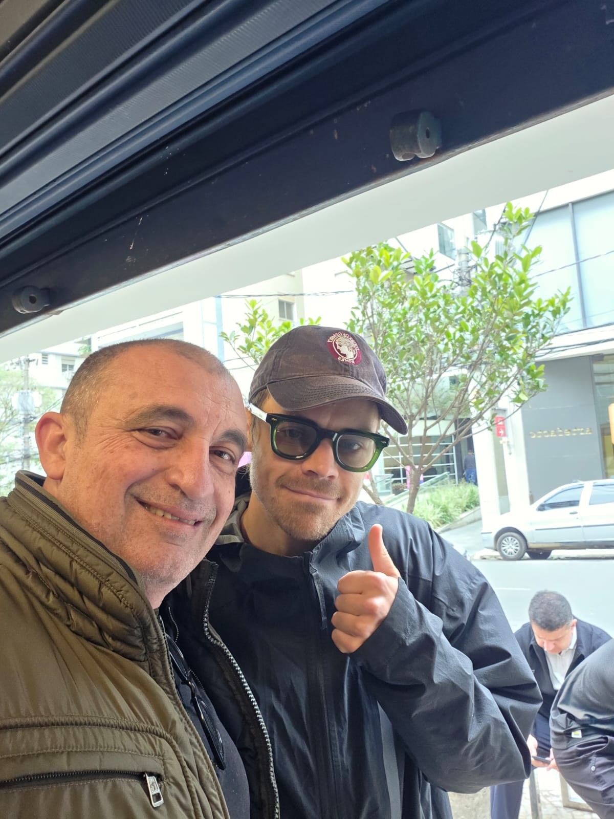 Dois homens sorrindo para a câmera, um com jaqueta verde-oliva e outro de boné, óculos e jaqueta preta, fazendo sinal de positivo. Ao fundo, uma rua com árvores e um carro branco estacionado