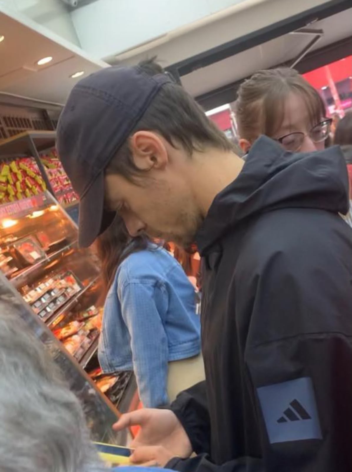 Homem de boné azul-marinho e jaqueta preta com logo Adidas no braço, olhando para baixo em um supermercado. Ao fundo, uma mulher de jaqueta jeans e outra de óculos. Prateleiras com produtos iluminados à esquerda.