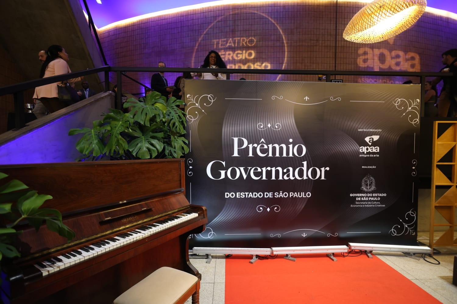 Um piano de madeira marrom com banco bege em primeiro plano, à esquerda. À direita, um banner preto com Prêmio Governador do Estado de São Paulo em branco. Um tapete vermelho se estende do banner. Ao fundo, pessoas sob luzes roxas, com Teatro Sérgio Cardoso visível na parede. Plantas verdes decoram o ambiente