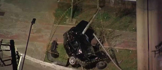 Carro preto capotado em área de grama ao lado de uma rua, com um poste de iluminação caído sobre ele e fitas de isolamento amarelas e pretas ao redor, à noite