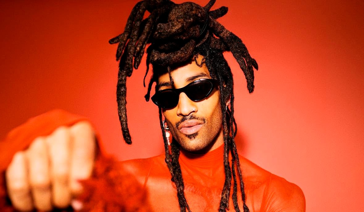 Homem negro com dreadlocks escuros, óculos de sol pretos e barba rala, vestindo uma blusa laranja translúcida, posa com o punho cerrado em primeiro plano, contra um fundo laranja vibrante