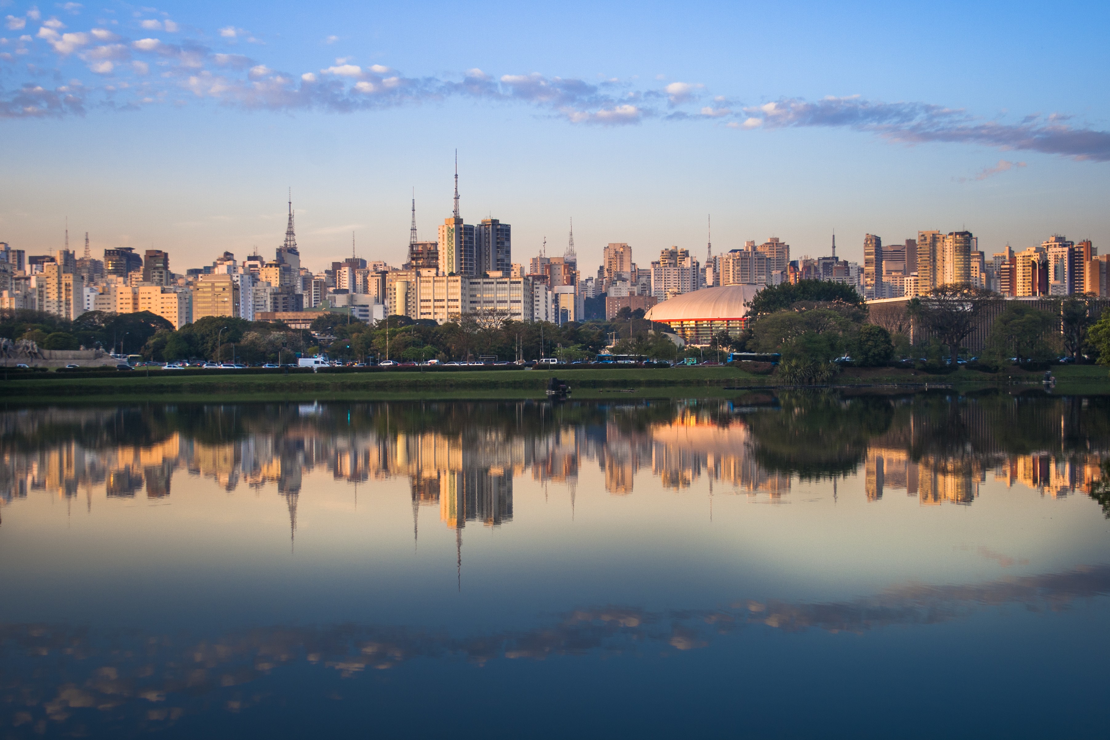 Panorama da cidade de São Paulo ao entardecer, com arranha-céus refletidos na superfície calma de um lago. O céu azul claro tem nuvens esparsas, e a luz dourada do sol ilumina os edifícios e a vegetação na margem.