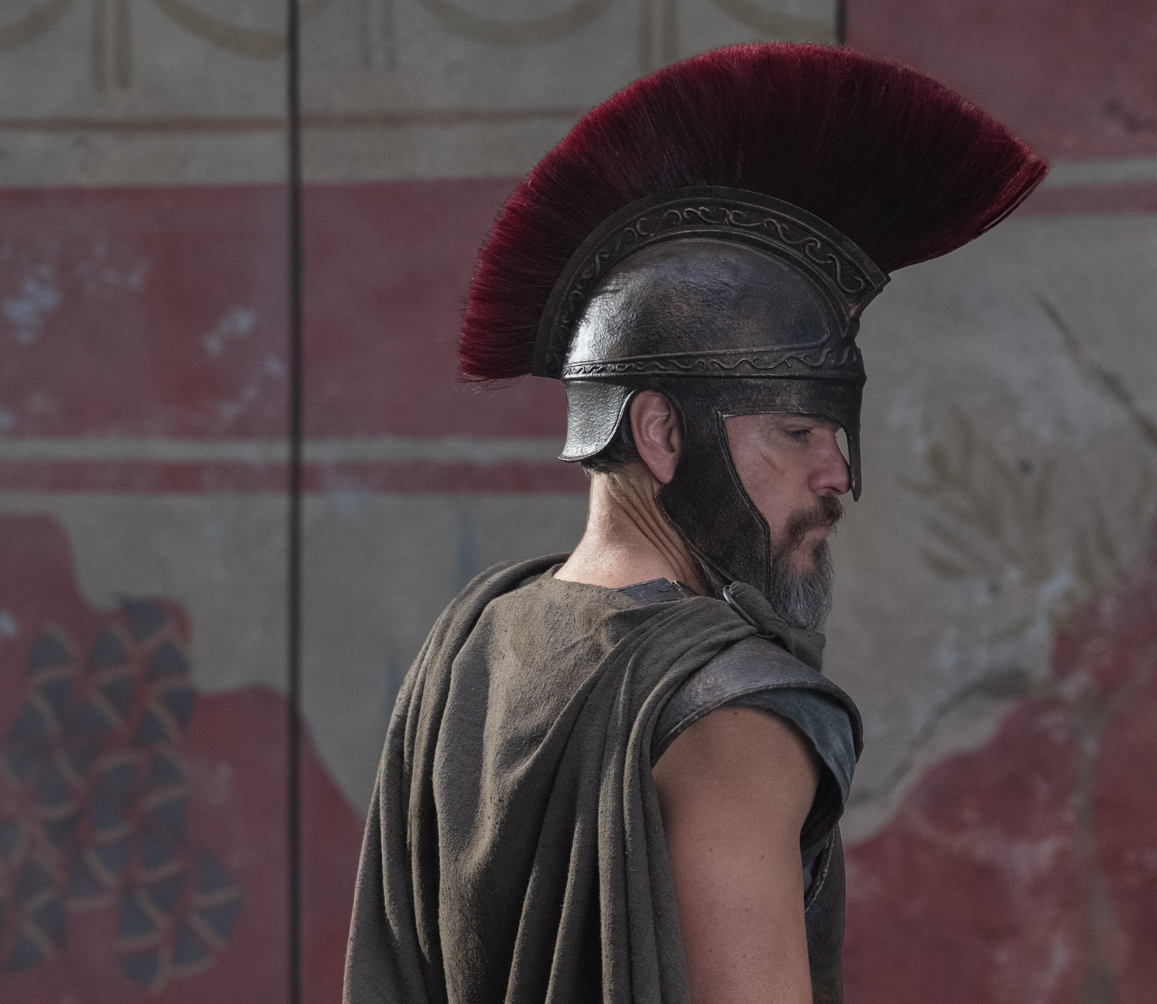 Homem barbudo de perfil, usando capacete espartano escuro com crista vermelha e capa marrom, olhando para baixo com expressão séria