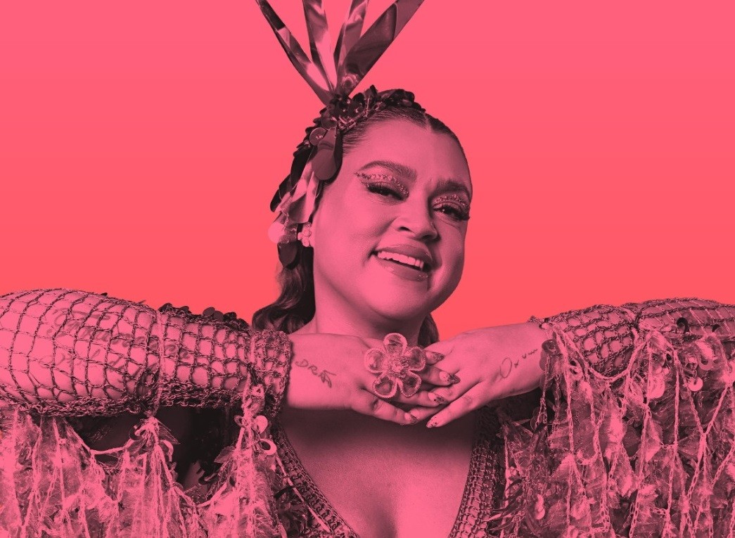 Mulher sorridente com maquiagem brilhante e adereço de cabeça com fitas, usando um vestido de rede e penas, com as mãos unidas sob o queixo e um anel de flor transparente, em fundo rosa