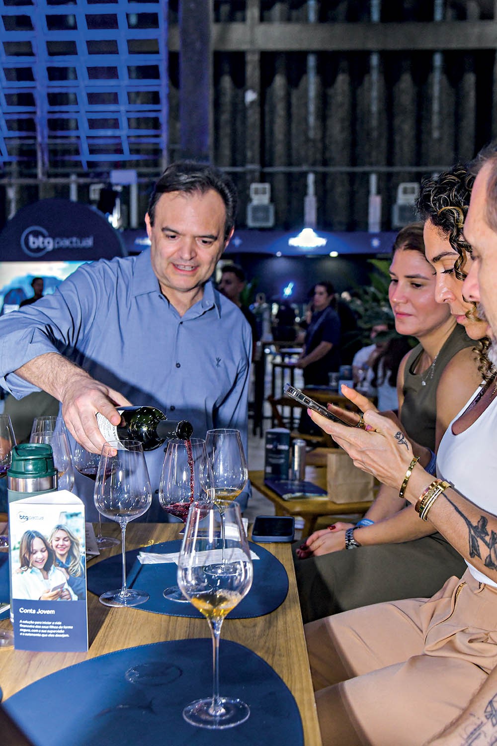 Homem de camisa azul serve vinho tinto em taças, enquanto três mulheres observam. Uma delas, com tatuagens e pulseiras douradas, segura um celular. Há um folheto Conta Jovem do BTG Pactual na mesa. O ambiente é um evento noturno com iluminação azulada