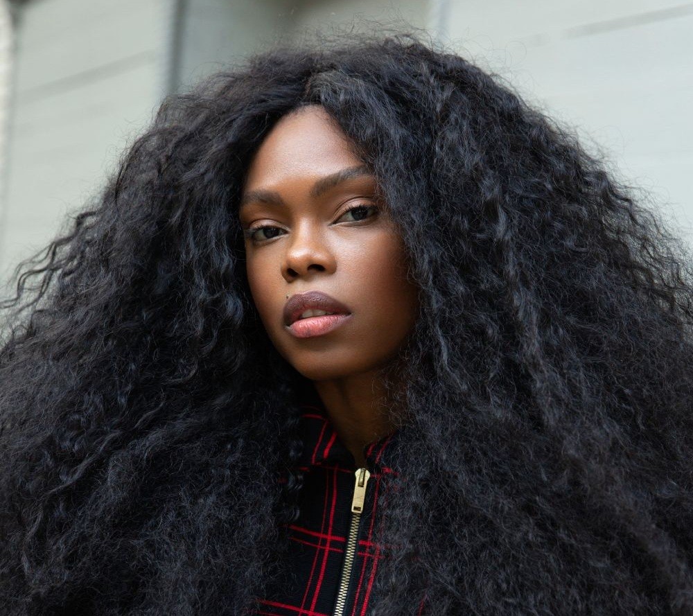 Mulher negra com cabelo crespo longo e volumoso, vestindo jaqueta xadrez preta e vermelha com zíper dourado, olhando diretamente para a câmera com expressão séria