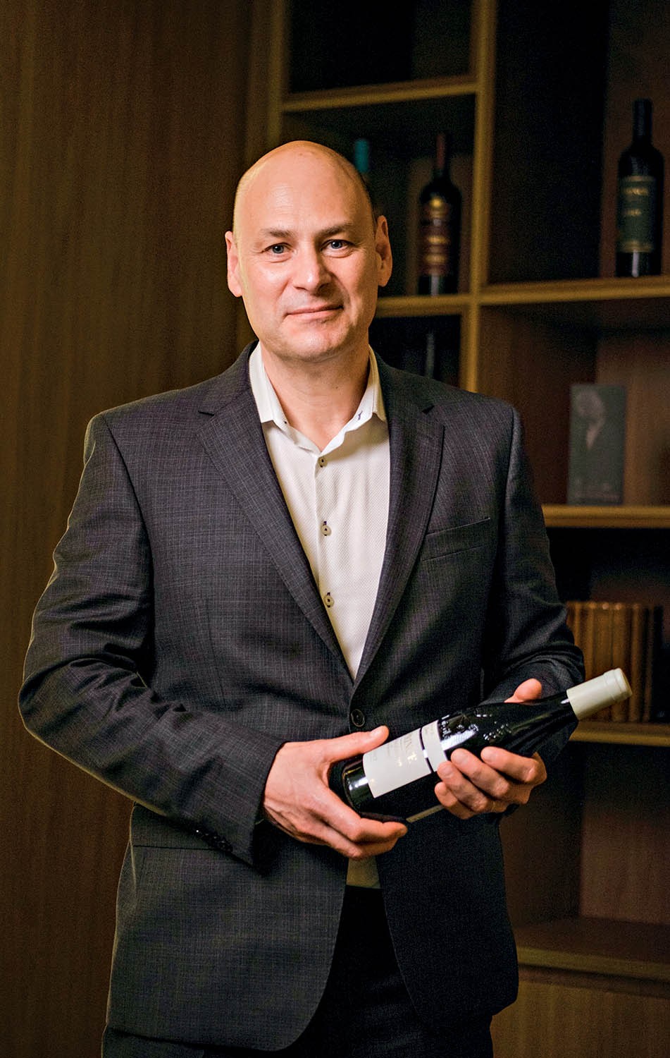 Homem calvo de meia-idade, vestindo terno cinza e camisa branca, segurando uma garrafa de vinho tinto com r&oacute;tulo branco. Ele sorri levemente para a c&acirc;mera, em frente a uma estante escura com garrafas e livros