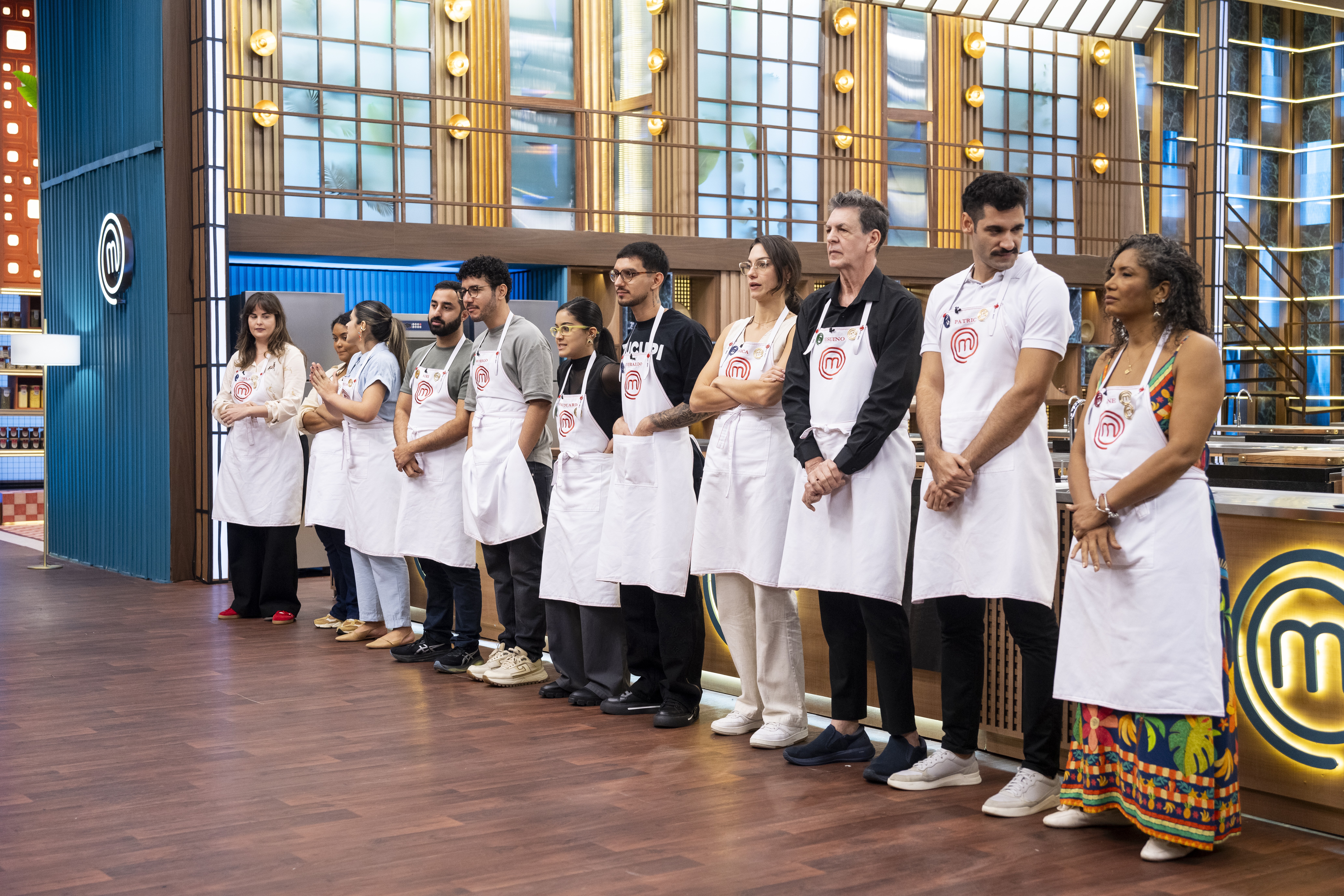 Um grupo de dez pessoas, incluindo homens e mulheres de diversas etnias, usando aventais brancos com o logo do MasterChef, estão em fila em um estúdio de televisão com paredes de madeira e detalhes em azul. Eles parecem atentos e sérios, aguardando instruções.