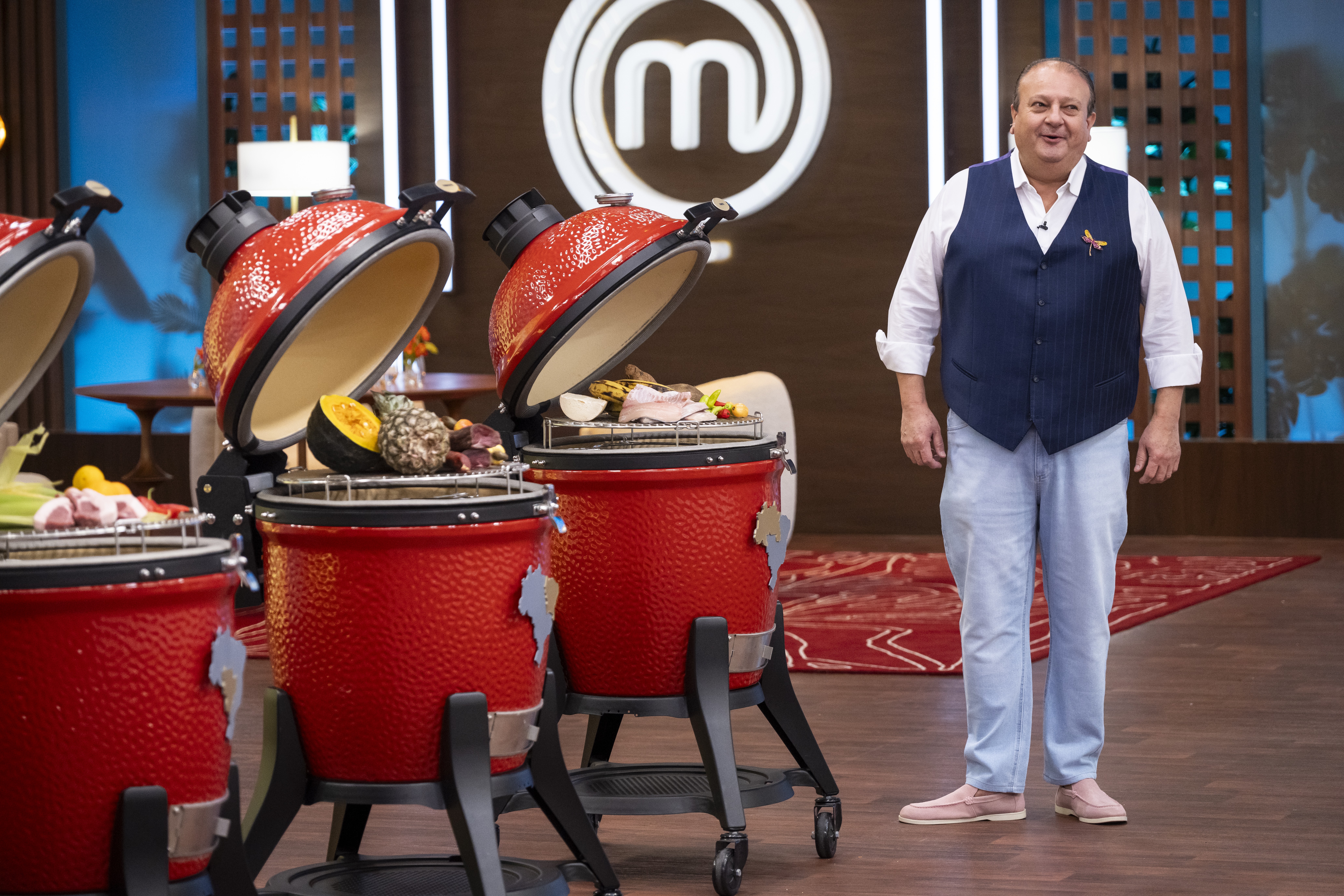 Homem sorridente de camisa branca, colete azul e calça clara, em pé ao lado de três churrasqueiras vermelhas com alimentos crus, no cenário do MasterChef