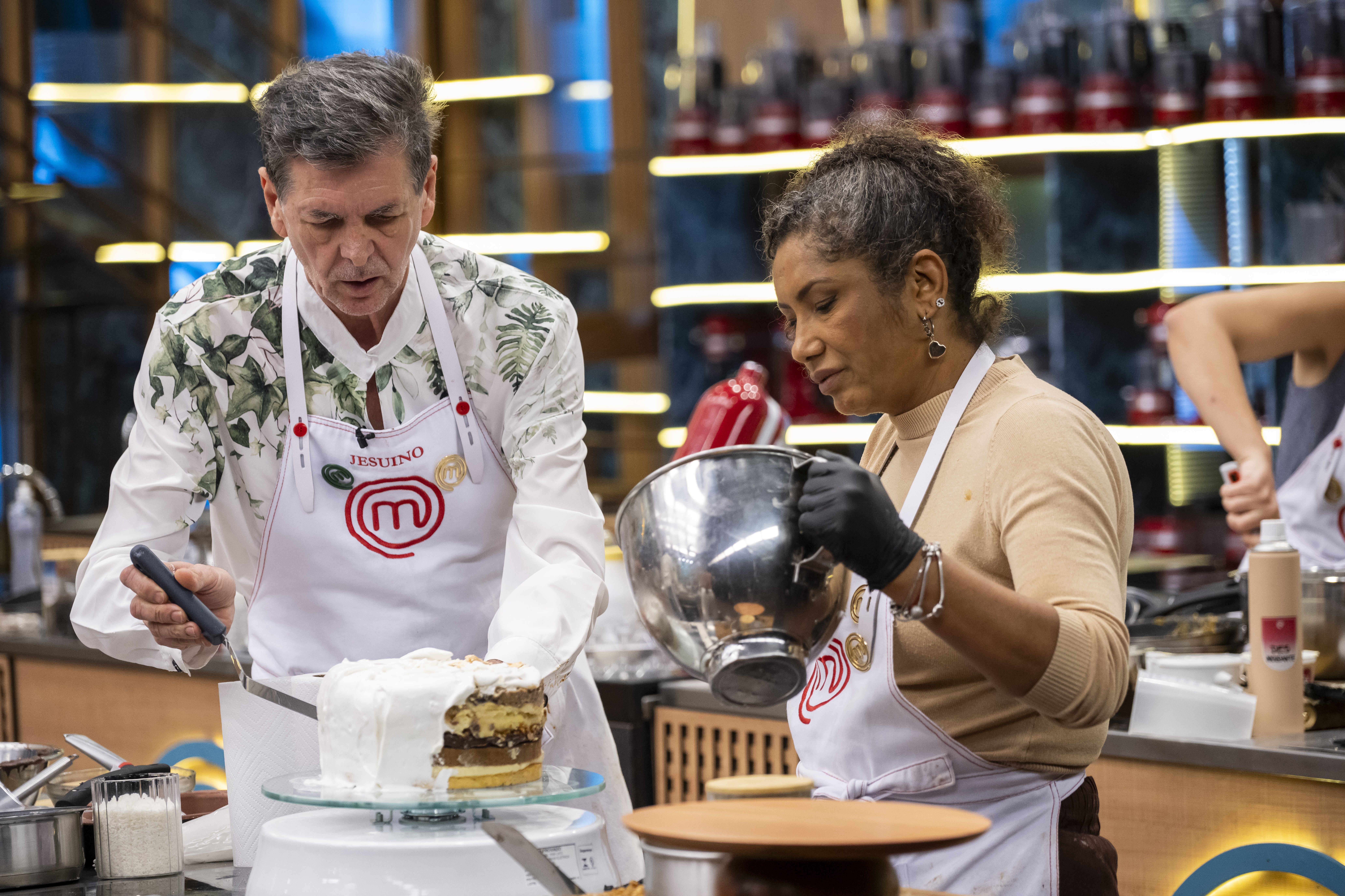 Um homem e uma mulher em aventais brancos de chef preparam um bolo. O homem, à esquerda, usa uma camisa estampada e espalha cobertura branca em um bolo de camadas. A mulher, à direita, com cabelo cacheado e luvas pretas, segura uma tigela de metal. Ambos estão concentrados na tarefa, em uma cozinha profissional com prateleiras iluminadas ao fundo