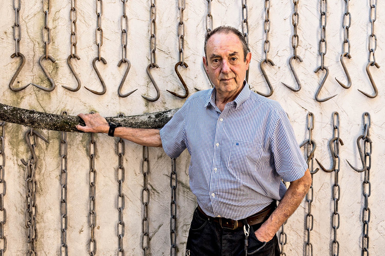 Homem idoso de camisa xadrez azul e branca, cal&ccedil;a escura e cinto marrom, encosta o bra&ccedil;o esquerdo em um tronco de &aacute;rvore. Ele est&aacute; em p&eacute;, olhando para frente, com um fundo de cor clara decorado com correntes e ganchos de metal pendurados