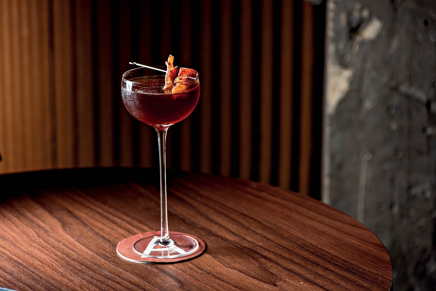 Coquetel vermelho escuro em ta&ccedil;a de haste fina, com guarni&ccedil;&atilde;o de bacon crocante espetado, sobre uma mesa de madeira escura e um porta-copos com a letra A. O fundo &eacute; desfocado, com listras verticais marrons e uma parede cinza texturizada