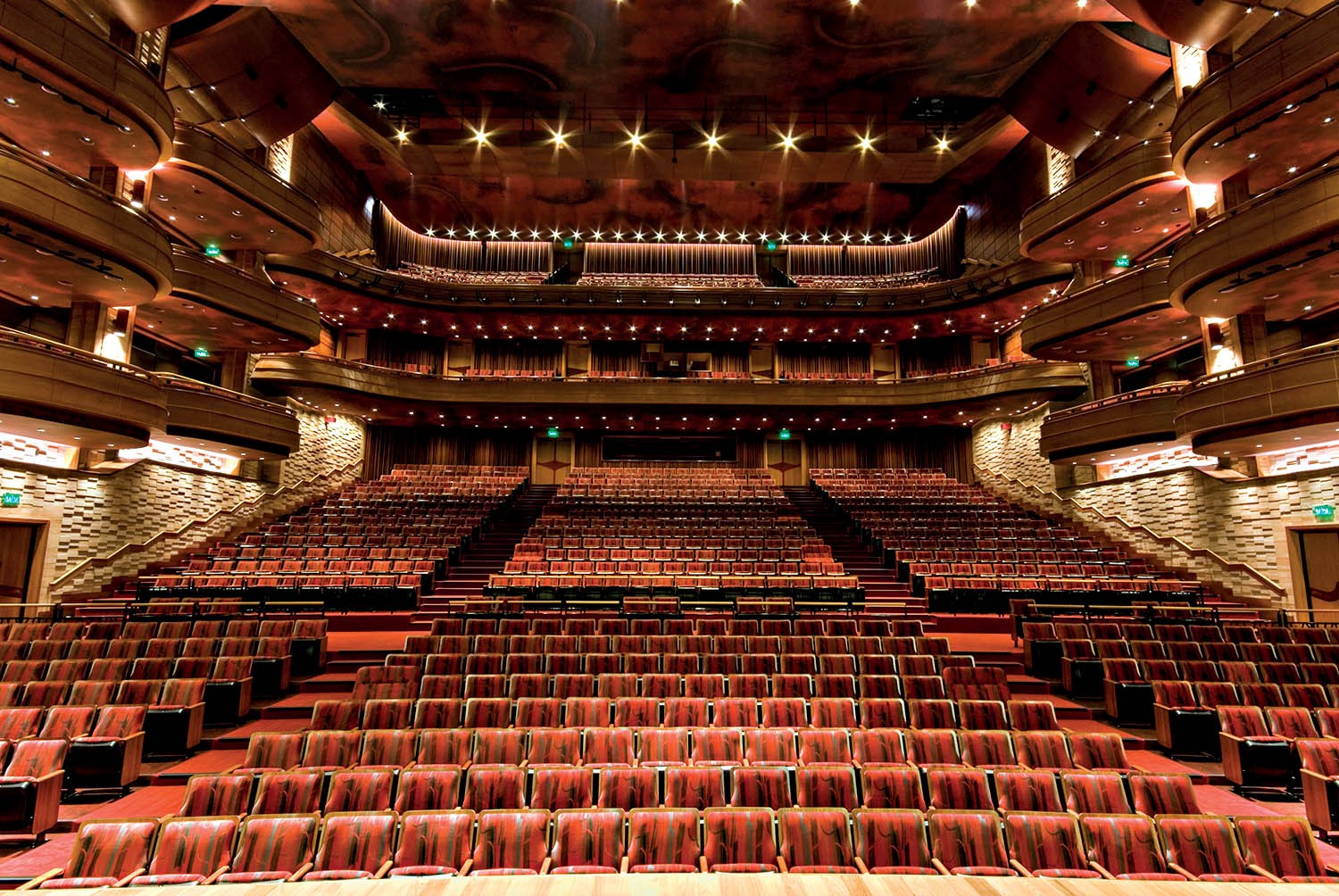 Interior de um grande teatro ou audit&oacute;rio com poltronas vermelhas vazias, dispostas em fileiras e v&aacute;rios n&iacute;veis de balc&otilde;es laterais. O teto abobadado tem ilumina&ccedil;&atilde;o pontual e detalhes dourados, criando uma atmosfera luxuosa