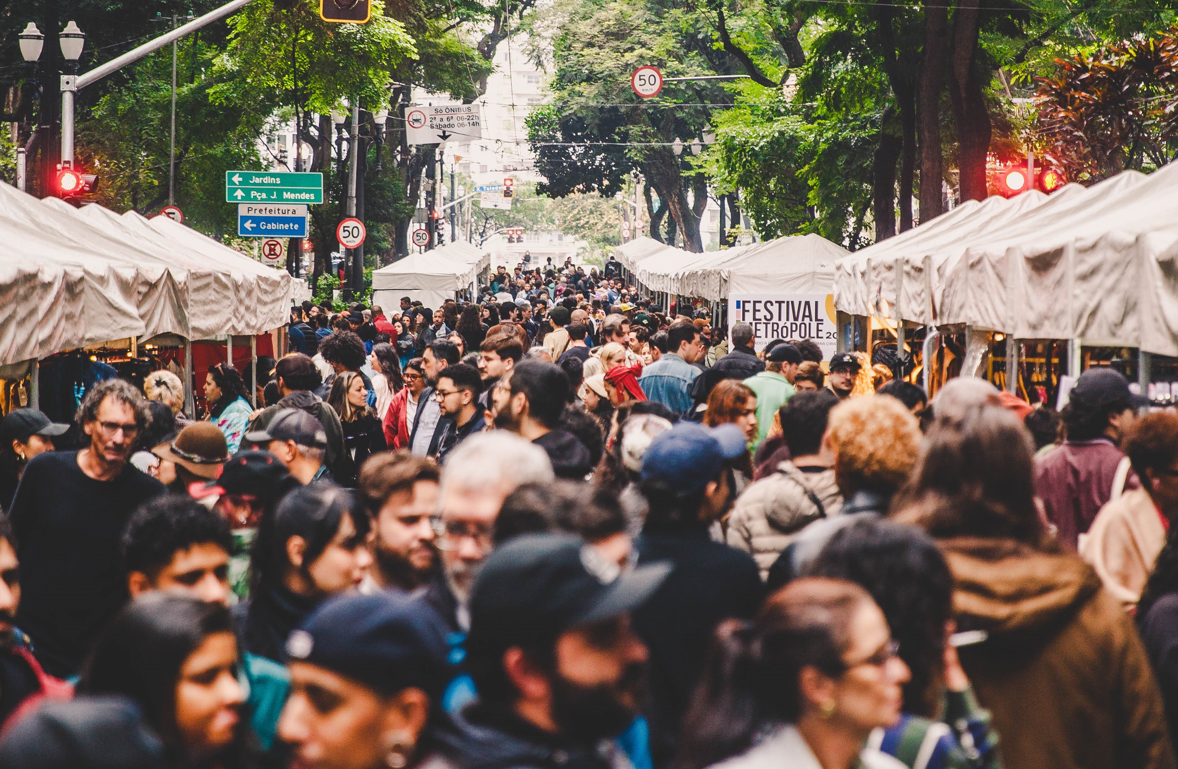Multidão de pessoas de diversas idades e etnias caminhando em rua arborizada, ladeada por barracas de feira com toldos brancos. Um semáforo vermelho e placas de trânsito são visíveis à esquerda, e uma barraca à direita exibe a inscrição FESTIVAL METROPOLE